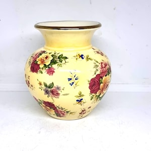 May include: A yellow ceramic vase with a floral design. The vase has a cream-coloured rim and is decorated with red, pink, and yellow roses, green leaves, and small blue flowers. The vase has a rounded body and a slightly flared neck.