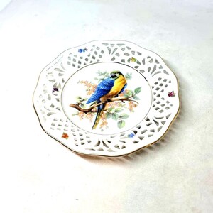 May include: White porcelain decorative plate with a gold rim and openwork design. The centre features a colourful illustration of a blue and yellow parrot on a flowering branch. A charming accent piece, approx. 20 cm in diameter.