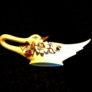 May include: A white ceramic swan ornament with a curved handle and a flared base. The swan has brown detailing on the beak and eyes, and is decorated with clusters of white and brown floral embellishments with gold accents.