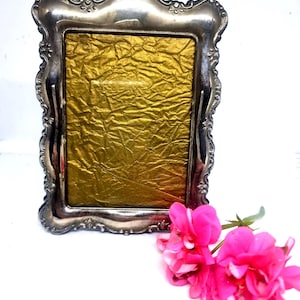 May include: An antique silver-coloured picture frame with a gold-coloured backing. The frame has an elaborate, decorative design. Two vibrant pink flowers lie in front. The frame is suitable for displaying photos or artwork.
