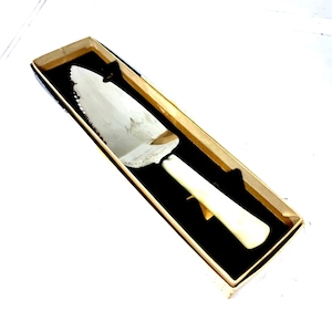 May include: A silver cake server with a serrated edge and a cream-coloured handle, presented in a rectangular box with a black interior and a light brown exterior. The server is designed for cutting and serving cake.