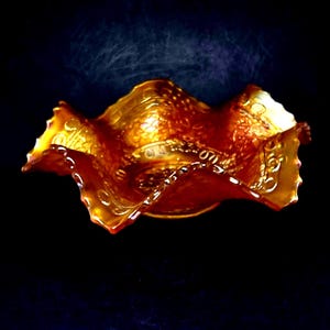 May include: An antique, iridescent orange glass bowl with a ruffled edge. The bowl has a textured surface with a pattern of dots and swirls. The bowl is set against a dark background.