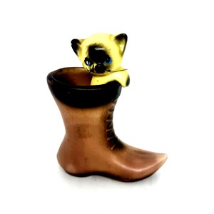 May include: A vintage ceramic figurine of a Siamese cat peeking out of a brown boot. The cat has a yellow face with black markings and blue eyes. The boot is brown with a black rim.