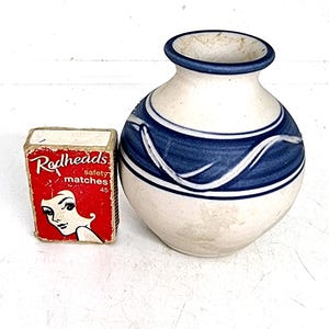 May include: A small, white ceramic vase with a blue rim and a blue and white decorative band. Beside the vase is a red matchbox with the text "Redheads" and a woman's illustration.