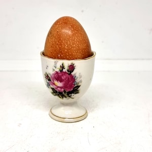 May include: A brown egg in a white porcelain egg cup. The egg cup has a floral design with a large pink rose and gold trim. The egg is light brown and has a textured surface. The egg cup is a vintage style.