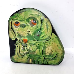 May include: A vintage, green dog-shaped tin with orange eyes and a small orange object in its paws. The dog is painted in shades of green and yellow with black outlines, set against a black background.