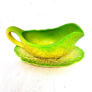 May include: A ceramic gravy boat and saucer set in shades of green and yellow. The gravy boat has a handle and a spout, and the saucer is round. The set has a textured, leaf-like design.