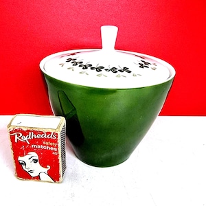 May include: A green ceramic sugar bowl with a white lid decorated with black shamrocks. A small box of "Redheads" safety matches is next to the bowl. The matchbox has a red background with a woman's face.
