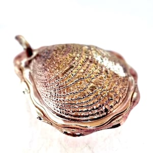 May include: A metallic pendant shaped like a clam shell with a textured, ridged surface in shades of bronze and copper. The pendant features a smooth, polished edge and a small loop at the top for attachment, showcasing a realistic shell design.