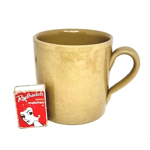 May include: A beige ceramic mug with a curved handle is positioned beside a red matchbox. The matchbox displays the text "Redheads" and an illustration of a woman with red hair. The mug has a speckled texture.