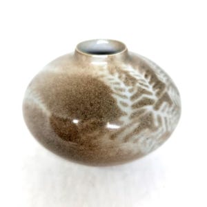 May include: A small, round ceramic vase with a speckled brown glaze. The vase has a white, fern-like pattern on its surface. The opening is a small, circular shape.