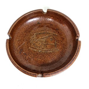 May include: Brown ceramic ashtray with a circular design. The ashtray has three notches for resting cigarettes and features an embossed image of a building with the text "Bendigo Pottery - Australia".