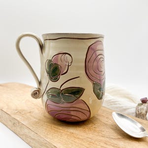 Può includere: Una tazza in ceramica con base color crema e un design floreale con rose rosa e verdi. La tazza ha un manico con una torsione unica.