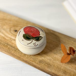Könnte beinhalten: Kleine, runde Schmuckdose aus cremefarbener Keramik mit einem roten Rosenmuster auf dem Deckel. Die Rose hat grüne Blätter und ist schwarz umrandet. Auf einer Holzoberfläche platziert.