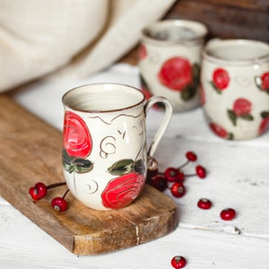 Könnte beinhalten: Drei weiße Keramikbecher mit roten und grünen Blumendesigns. Die Becher stehen auf einem Holzbrett und einer weißen Oberfläche. Um die Becher herum liegen rote Beeren verstreut.