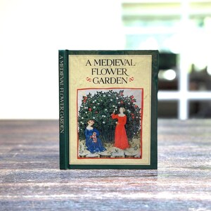 May include: A hardcover book titled "A Medieval Flower Garden." The cover features an illustration of two figures in medieval attire, one in a red dress and the other in blue, amidst a flowering bush. The book's spine is green.