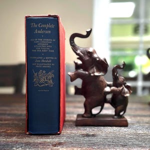May include: A hardcover book titled "The Complete Andersen" with blue cover and red spine, next to a bronze elephant figurine. The book's cover features gold lettering and illustrations. The elephant sculpture depicts a parent and child.