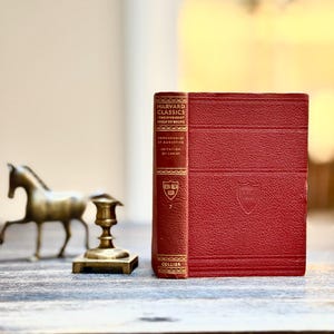 May include: A red hardcover book from the Harvard Classics series, titled "Confessions of St. Augustine" and "Imitation of Christ." The book has gold lettering and a decorative spine. A small brass horse figurine and candle holder are in the background.