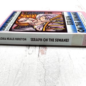 Puede incluir: Un libro de tapa dura titulado "Seraph on the Suwanee" de Zora Neale Hurston. El libro tiene una cubierta rosa y una ilustraci&oacute;n colorida en la portada. El libro est&aacute; sobre una superficie blanca.