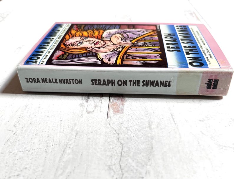 Puede incluir: Un libro de tapa dura titulado "Seraph on the Suwanee" de Zora Neale Hurston. El libro tiene una cubierta rosa y una ilustraci&oacute;n colorida en la portada. El libro est&aacute; sobre una superficie blanca.