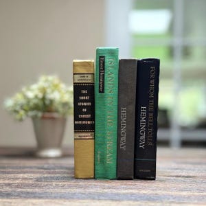 May include: Four hardcover books by Ernest Hemingway stand upright on a wooden surface. The book spines are in various colors: gold, green, gray, and navy blue. Titles include "The Short Stories", "Islands in the Stream", and "For Whom the Bell Tolls".