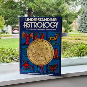 May include: A hardcover book titled "Understanding Astrology" with a dark blue cover. The cover features a gold-colored circular design and illustrations of zodiac symbols. The text "The Influence of the Stars on You and Others" is also visible.