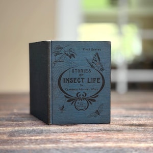 May include: A vintage blue book titled "Stories of Insect Life" by Clarence Moores Weed. The cover features illustrations of insects, a spider web, and a butterfly. The book is from the "First Series" and published by Ginn and Company.