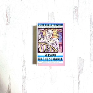 Puede incluir: Un libro de bolsillo titulado "Seraph on the Suwanee" de Zora Neale Hurston. La portada presenta una ilustraci&oacute;n de una mujer con cabello rubio, apoyando la barbilla en su mano. El libro tiene un borde rosa.