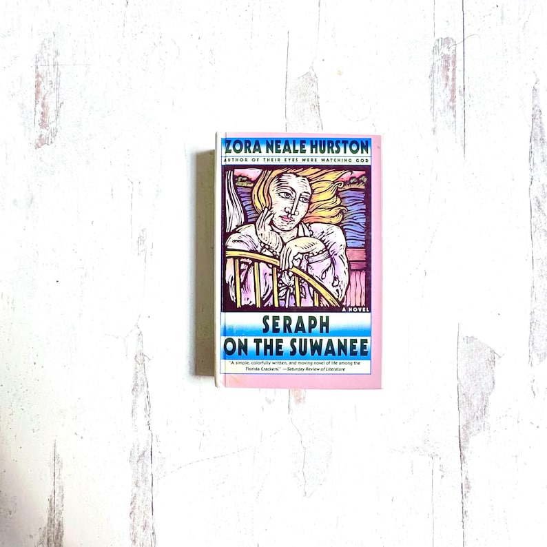 Puede incluir: Un libro de bolsillo titulado "Seraph on the Suwanee" de Zora Neale Hurston. La portada presenta una ilustraci&oacute;n de una mujer con cabello rubio, apoyando la barbilla en su mano. El libro tiene un borde rosa.