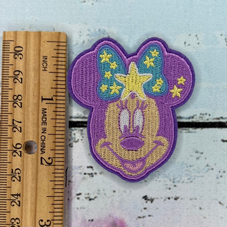 K&ouml;nnte beinhalten: Lilafarbener gestickter B&uuml;gel-Patch mit einer Comic-Maus mit einem gelben Stern und einer blauen Schleife mit gelben Sternen.