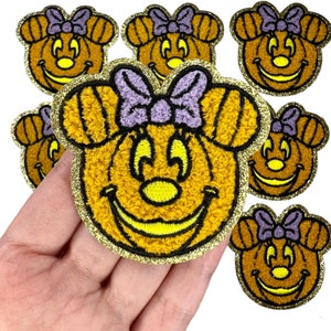 May include: Set of seven iron-on patches featuring a cartoon pumpkin with a bow. The pumpkin is orange with a yellow smile and eyes. The bow is purple. The patches are surrounded by a gold glitter trim.