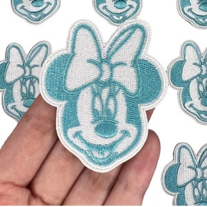 May include: Embroidered patches featuring a cartoon character's face with a large bow. The patches are light blue and white, with a detailed stitched design. The patches are likely for clothing or accessories.