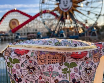 Puede incluir: Una riñonera blanca con un estampado colorido que presenta a Mickey Mouse, una noria y otras atracciones del parque de Disney. El estampado también incluye el texto "Tiki Juice Cafe".