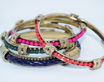 Può includere: Set di quattro braccialetti dorati con accenti di perline colorate. I braccialetti sono impilati uno sopra l'altro, con il braccialetto superiore con perline rosa, il secondo braccialetto con perline verdi, il terzo braccialetto con perline rosse e il braccialetto inferiore con un cinturino in pelle blu scuro.