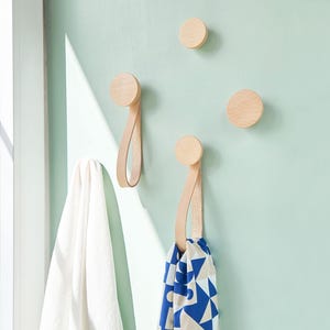以下が含まれることがあります： 革のループが付いた木製壁掛けフックが3つ。2つのフックには、白いタオルと青と白の模様のストールがかけられています。フックは薄い緑色の壁に取り付けられています。