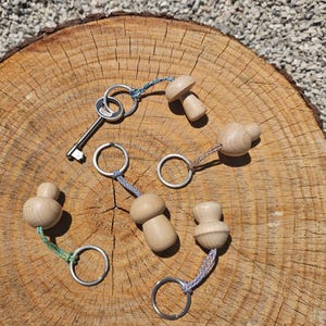 Op de afbeelding: Vier sleutelhangers met gladde, lichtgekleurde houten kralen in de vorm van paddenstoelen, bevestigd aan zilverkleurige metalen ringen met kleurrijke gevlochten koorden. Eén sleutelhanger bevat een zilverkleurige metalen sleutel.