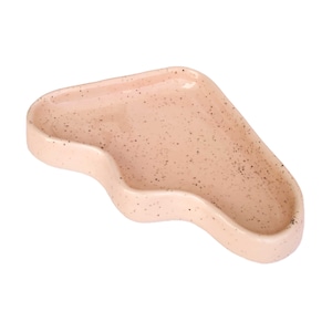 May include: A speckled, light pink ceramic spoon rest with a unique, wavy shape. The dish has a shallow, curved design with a slightly raised edge. The surface is dotted with small, dark brown specks.