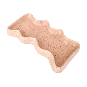 May include: A light pink ceramic serving dish with a wavy, organic shape. The dish has a speckled texture.