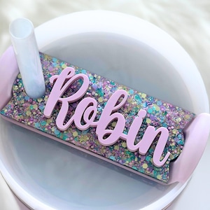 May include: A light purple glittery tumbler with the name "Robin" in cursive. The tumbler has a light purple handle and a clear straw. The glitter is a mix of colors, including purple, blue, and green.