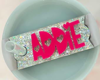 May include: A pink glitter tumbler with a pink name tag that says "ADDIE" with hearts. The name tag is attached to the tumbler with a clear plastic strap.