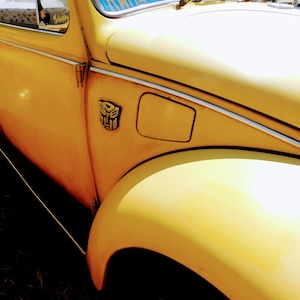 May include: Close-up of a yellow vintage car, featuring a rounded front fender and a side panel with a silver emblem. The car's body has a glossy finish, with a chrome trim along the side. The image is taken in daylight.