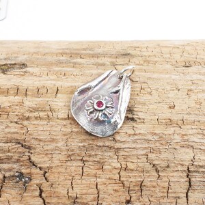May include: A silver pendant with a textured, organic shape featuring a raised flower design with a red gemstone in the center.