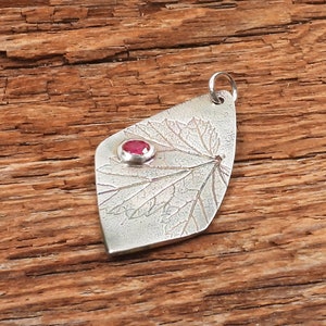May include: A silver pendant with a detailed leaf design and a small red gemstone in the center. The pendant is on a wooden surface.