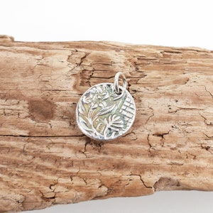 May include: A silver pendant with a detailed floral design. The pendant is round and has a textured surface. The flowers are stylized and have a green and white color scheme.