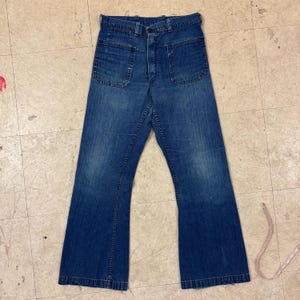 Può includere: Jeans blu in denim con ampie tasche frontali e un design a zampa d'elefante. I jeans hanno una chiusura con bottone e cerniera e mostrano segni di usura. I jeans sono stesi su una superficie chiara.