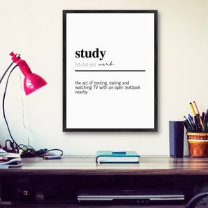 Puede incluir: Una impresión enmarcada en blanco y negro con la palabra "study" definida como "el acto de enviar mensajes de texto, comer y ver televisión con un libro de texto abierto cerca". La impresión está colgada en una pared blanca sobre un escritorio con una lámpara, una planta en maceta y una silla.