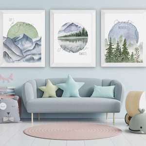 May include: Three framed watercolor art prints with nature scenes. The prints feature mountains, a lake, and trees, with the phrases "Let's Explore," "Adventure Awaits," and "Create Memories." The art is displayed above a gray sofa with star-shaped pillows.