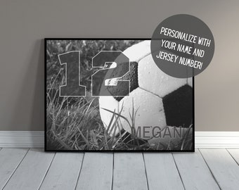 May include: Black and white framed print featuring a soccer ball in grass, with the number 12 overlaid. The name "Megan" is printed below the ball. The print is in a black frame.