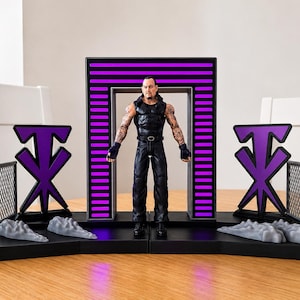 May include: A plastic action figure of a wrestler with tattoos, wearing a black sleeveless top and trousers, standing in a black and purple arched entrance display. The set includes two purple logo signs, two grey chain-link fences, and four grey rock pieces.
