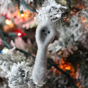May include: A gray felt ornament shaped like a ghost with a simple face, hanging on a snowy Christmas tree with twinkling lights.